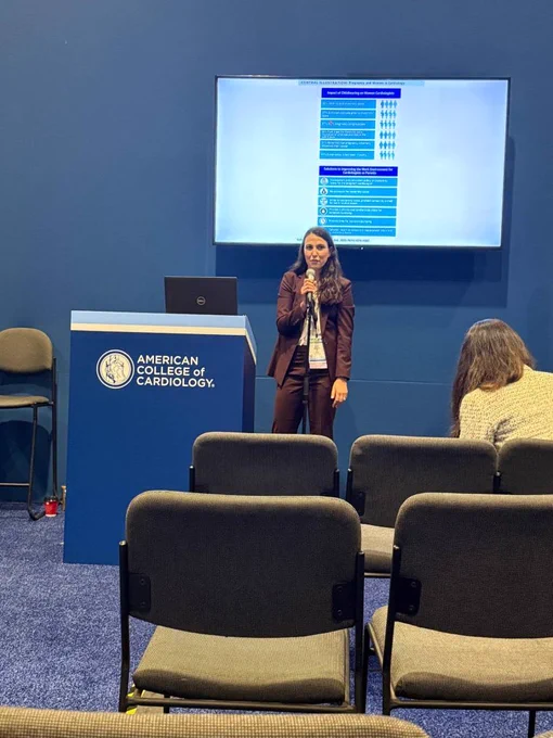 A presenter stands with a microphone beside an American College of Cardiology podium in a small session room, speaking in front of a wall-mounted screen while audience chairs fill the foreground.