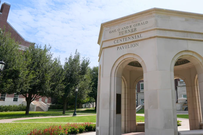 The image shows the Gail O. and R. Gerald Turner Centennial Pavilion, an architectural structure with arches, located on a grassy area surrounded by trees and flowers. A brick building is visible in the background.