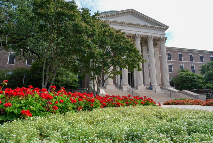 The image shows the exterior of Dallas Hall at SMU, a large classical-style building with tall white columns at the entrance, surrounded by neatly landscaped gardens featuring red and white flowers beneath a partly cloudy sky.