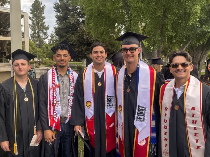 Five Stan State graduates pose together outdoors in caps and gowns wearing various honor stoles, cords, and medals during commencement celebrations.