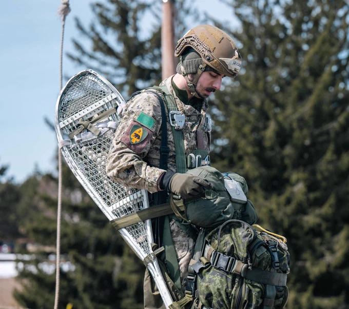 Soldier in full gear with parachute pack and snowshoes secured to a rucksack checks equipment during outdoor training.