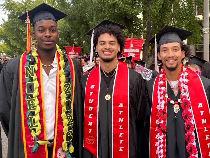 Three Stan State graduates stand together in caps and gowns wearing decorated leis and red “Student Athlete” stoles while smiling for a commencement photo.