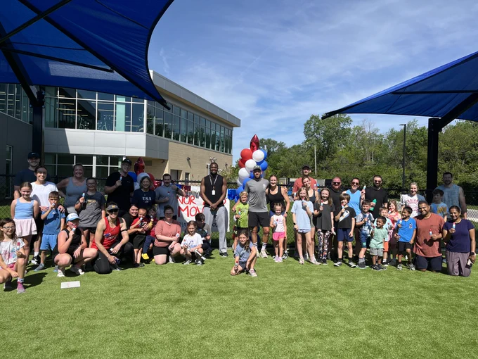 A large group of people, including children and adults, are gathered on a grassy area outside of the Fishers Community Center. They are posing for a photo under blue shade structures. In the background, there are colorful balloons. The sky is clear and sunny.