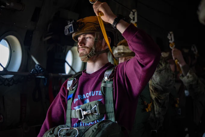 Paratrooper inside a military aircraft attaches a static line to an overhead cable while conducting pre-jump checks; other personnel prepare in the background.