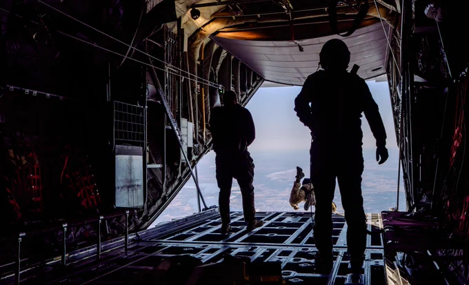 Silhouettes of paratroopers at the open ramp of a military aircraft as one exits and jumps over a landscape far below.