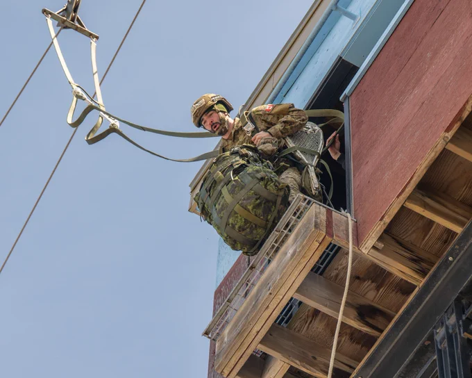 Paratrooper exits a training tower while connected to overhead rigging, carrying full gear including a rucksack and snowshoes during airborne training.