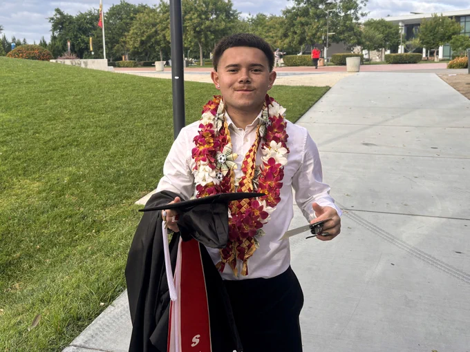 A Stan State graduate wearing flower leis and holding graduation regalia smiles while standing on a campus walkway after commencement.