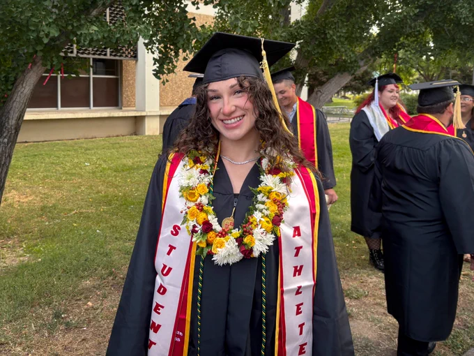 A smiling Stan State graduate in a black cap and gown poses outdoors wearing multiple flower leis and a red-and-white “Student Athlete” stole during commencement.