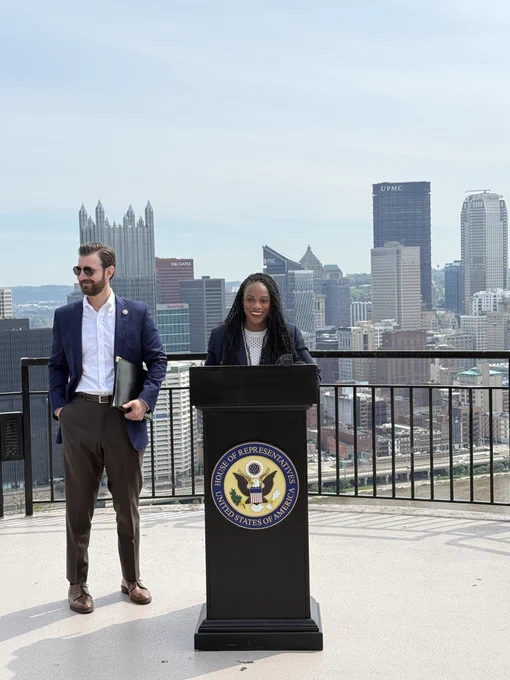 Rep. Summer Lee stands behind a podium and is smiling. To her left is Rep. Chris DeLuzio. Behind them is the Pittsburgh skyline.