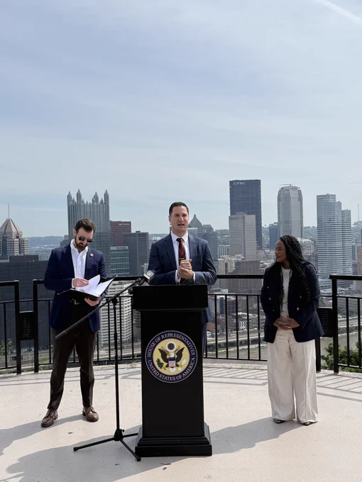 Kevin Zedack of Appalachian Voices stands behind a podium and is speaking into a mic. To his right is Congresswoman Summer Lee. To his left is Congressman Chris DeLuzio. They are standing at an overlook with the Pittsburgh skyline behind them.