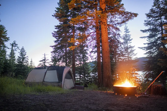 A glowing campfire beside a tent in a forest clearing at dusk with tall pine trees and a lake in the background.