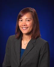 Professional portrait of Embry-Riddle Assistant Professor of Mathematics Dr. Angelynn Alvarez wearing a dark gray blazer, blue top, and necklace against a deep blue background.