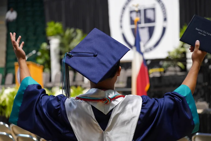 Two graduates in graduation gowns and caps, hands raised excitedly in front of a university banner.