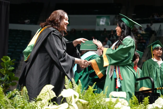 Two graduates in green and black robes shaking hands at an outdoor graduation ceremony.