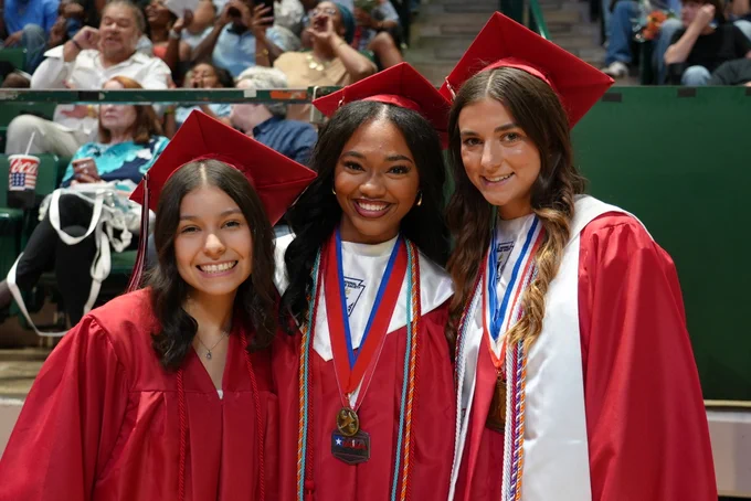 Three smiling female graduates in red caps and gowns standing together.
