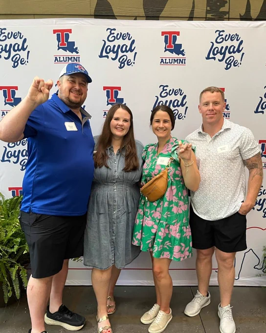 Four people stand in front of a Louisiana Tech alumni banner. Two make a bulldog symbol with one hand