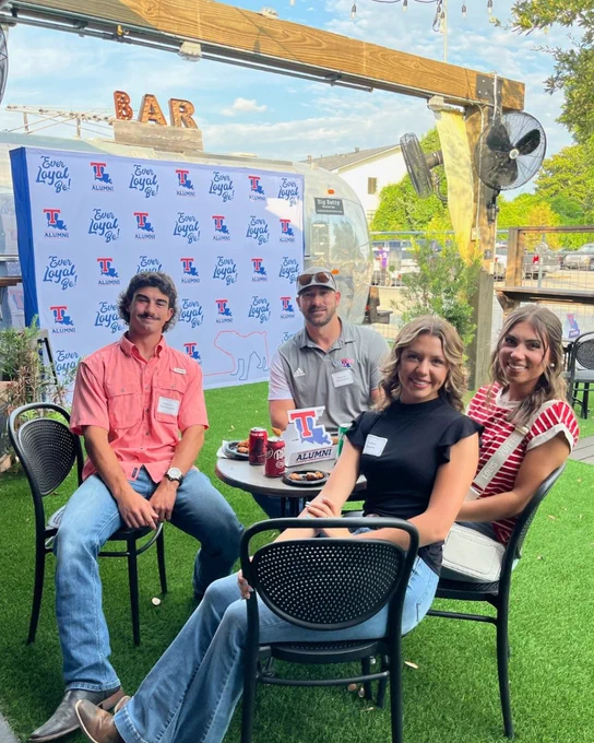 Four people sit at an outdoor table with a Louisiana Tech alumni sign on it