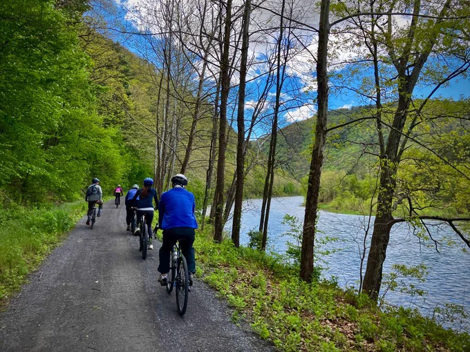 people riding on a bike trail along the river. There is bright green vegetation on both sides of the trail. 