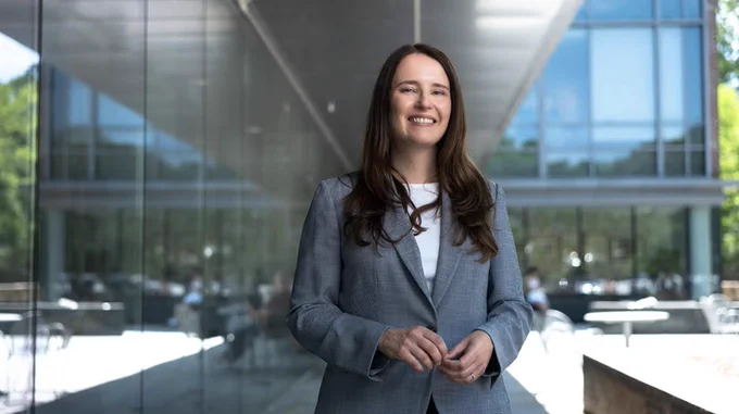 the image shows someone smiling and standing outdoors in front of a modern glass building.