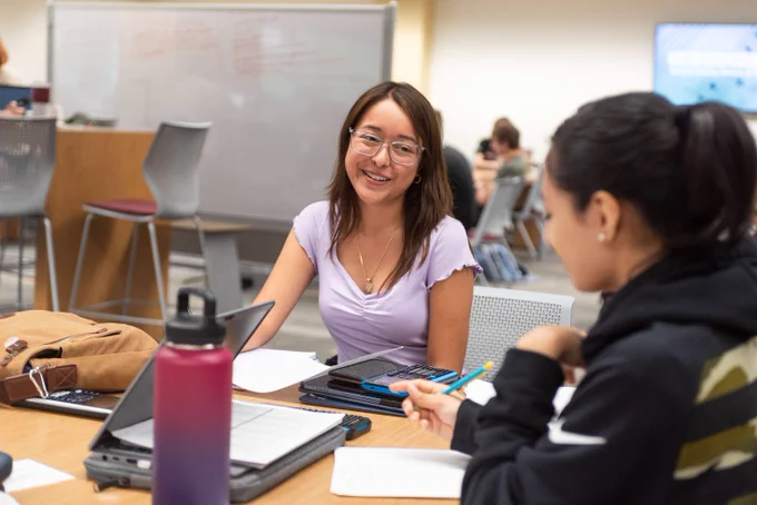 Two students engaged in a discussion at a classroom table with laptops and notebooks open.