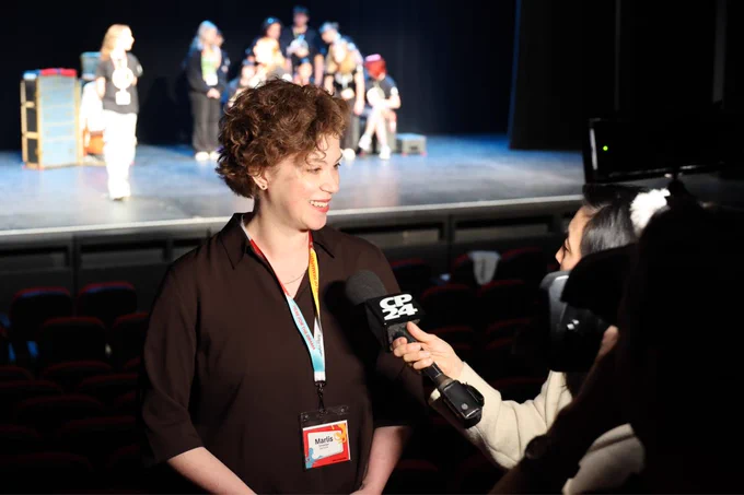 A person wearing a name badge is being interviewed by a CP24 reporter in an auditorium. In the background, a group of individuals are gathered on a stage.