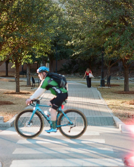 Cyclist wearing a helmet and green jersey rides a blue bike across a pedestrian crosswalk in a park with trees and benches.
