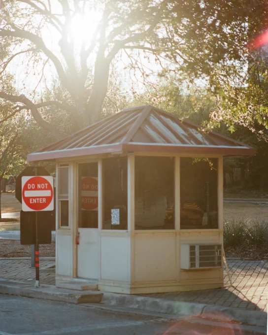 Small guard booth with a rusty metal roof beside a 'Do Not Enter' sign under bright sunlight filtering through tree branches.