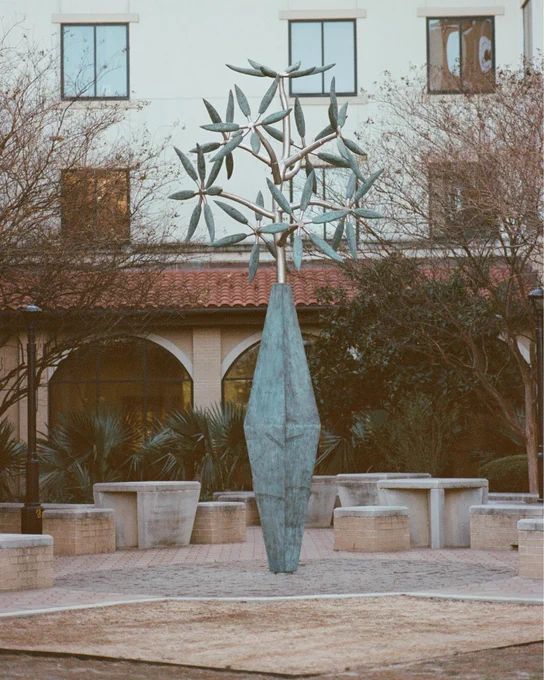Tall abstract metal sculpture resembling a tree with multiple leaf-like shapes in a courtyard with benches and a building backdrop.
