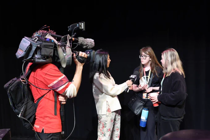 A person holding a large video camera is filming a discussion between three individuals. 