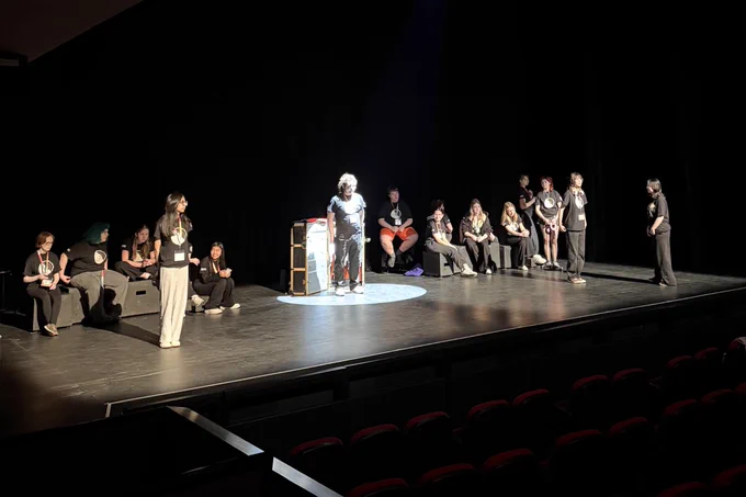 A group of performers are on a dimly lit stage, with one person standing in a spotlight near the center. 