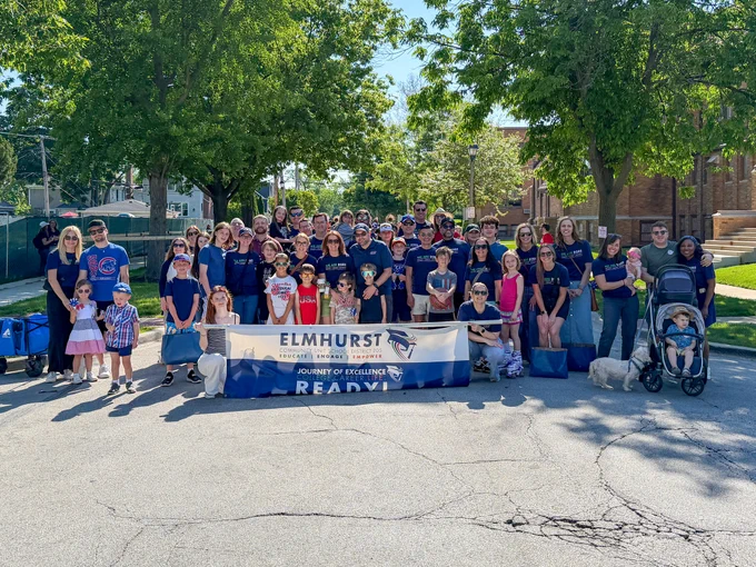 A large group of people including children and adults stand together outdoors, holding a banner for Elmhurst Community Unit School District 205. They appear cheerful and are posing for the camera on a sunny day.