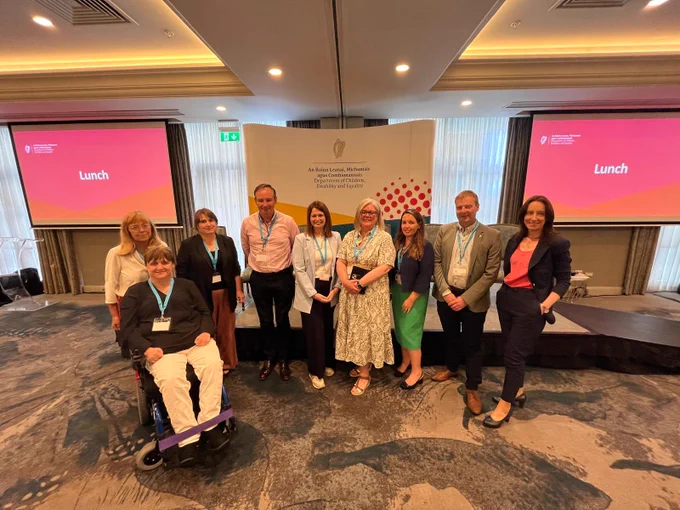 A group of nine people pose for a photo at the Department of Children, Disability and Equality stakeholder forum at the Hilton Hotel, Dublin. One person in the group uses a powered wheelchair. Behind them is a branded backdrop showing the Department's name in Irish and English, flanked by two large screens displaying a colourful event slide. The group are wearing lanyards and name badges.