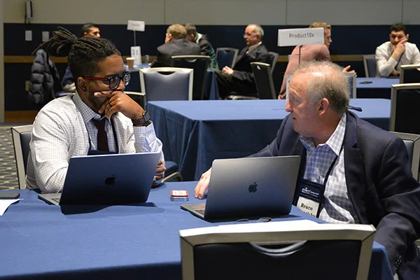 Two men in business attire engage in a focused discussion across a table with laptops during a professional networking event.