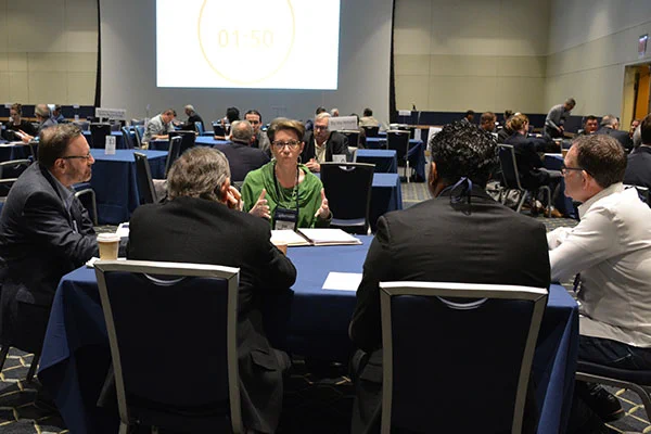 Group of professionals engaged in a focused discussion around a conference table in a large meeting room with a timer projected on the screen.