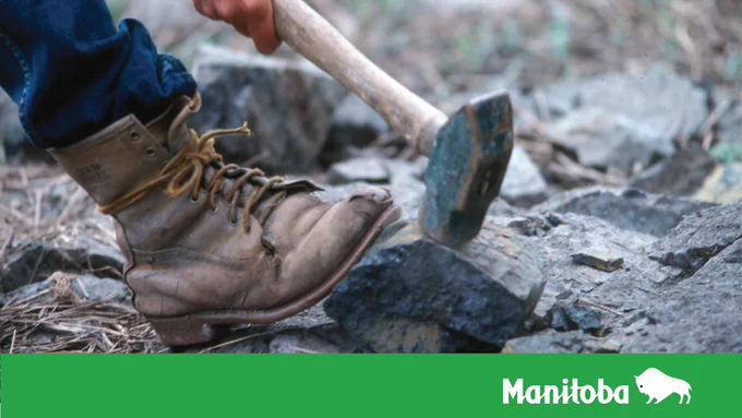 Photo of a geologist's boot resting against a rock while holding a rock hammer on top of the rock as if striking it.