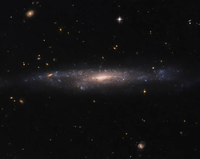 A galaxy extends across the center of the image, with a yellow core and surrounded by blue stars and brown dust. Against the black background of space, foreground stars and more distant galaxies shine.