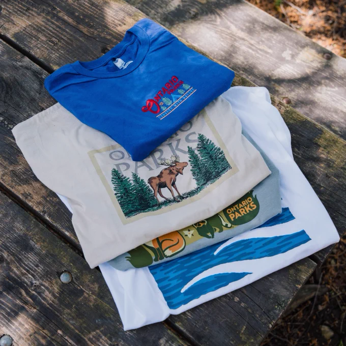 Ontario Parks blue t-shirt with logo on the front, moose and trees tote bag, and other merchandise on a picnic table.