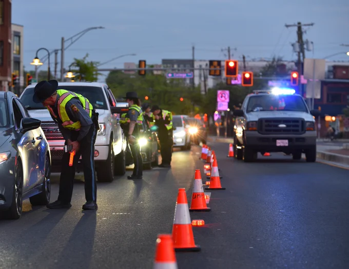 A line of cars are stopped in a lane of traffic with a police officer standing at the driver's side window of each vehicle as they have entered a sobriety checkpoint.  