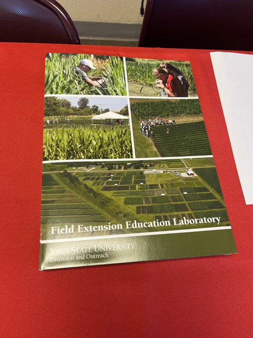 A green folder with images of crop fields and people learning at events on a table with a red tablecloth.