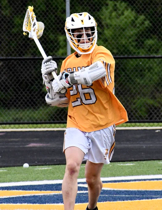Adams lacrosse player wearing  number 26 prepares to pass the ball on a field with a black fence in the background.