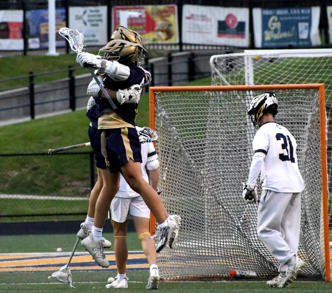 Two Stoney Creek lacrosse players in dark uniforms jump in celebration near the goal while an opposing goalie in white watches.