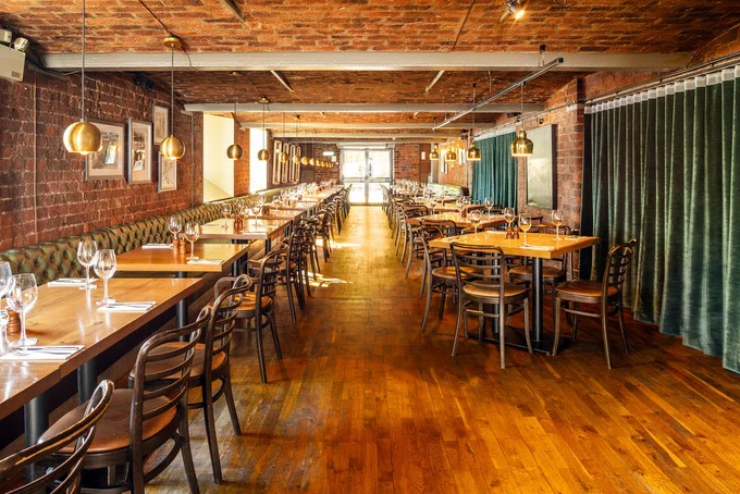 Spacious restaurant interior with exposed brick walls, wooden floors, long communal tables set with glassware, green banqueting seating and warm pendant lights.