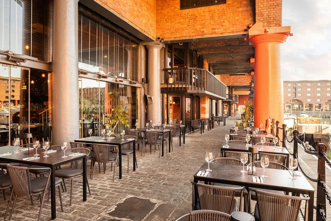 Outdoor waterside area with neatly set tables and chairs beneath a covered brick colonnade, overlooking a marina and historic dock building.