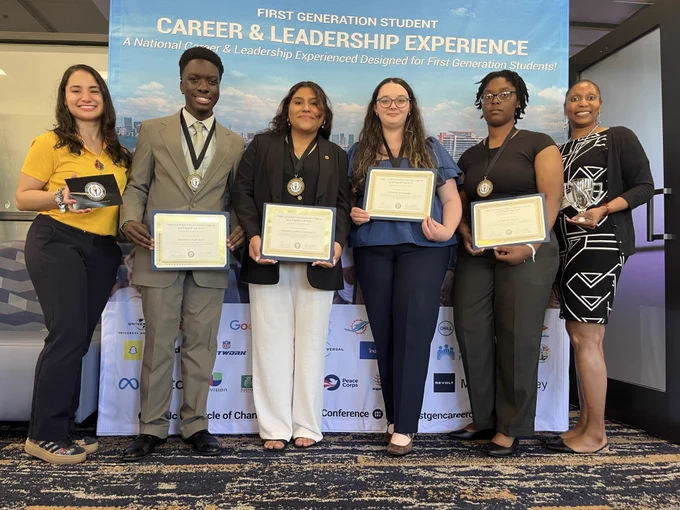 A group of individuals stand proudly in front of a banner that reads "First Generation Student Career & Leadership Experience." They hold certificates and wear medals around their necks. The setting appears to be an indoor event.