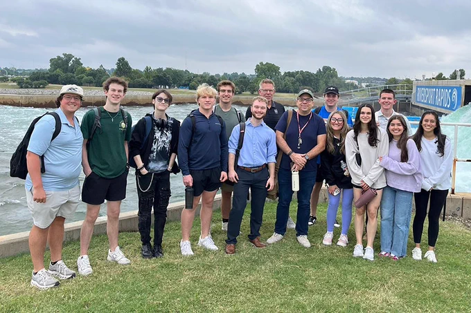 A group of people are standing on grass near a body of water. They are casually dressed and smiling at the camera. University of Oklahoma students taking the school's Applied Social Innovation course combined their entrepreneurship and engineering skills to help protect the Oklahoma River for the upcoming Summer Olympics. Photo by Bruno Teles.