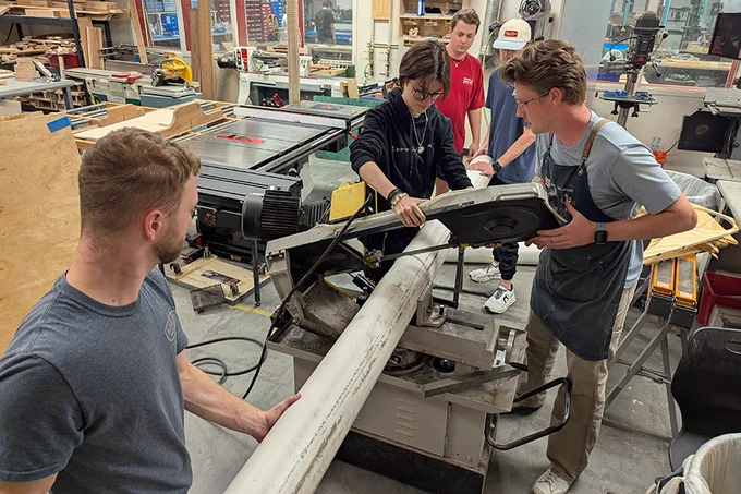 A group of people work collaboratively in a workshop, focused on a machine with a large cylindrical object. The setting is industrial with tables, tools, and machines in the background. One person adjusts the machine while others assist, and the environment suggests teamwork and hands‑on technical activity.
