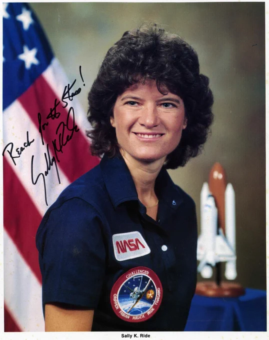 A person wearing a NASA uniform with the Space Shuttle emblem poses in front of a U.S. flag and a model of a space shuttle. There is an autograph and a message reading, "Reach for the stars!" The name "Sally K. Ride" is noted below the image.