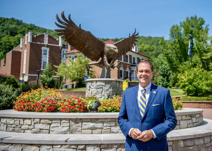 Morehead State University President Dr. Jay Morgan standing in front of of MSU’s large Centennial Eagle sculpture and double layered stone base. A few campus buildings and trees are in the background.