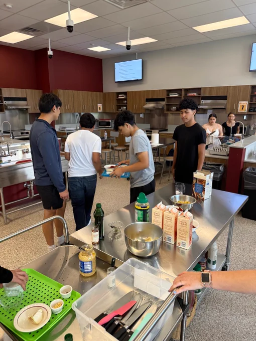 A group of people in a kitchen setting, with cooking supplies and appliances visible. Appears to be a culinary or teaching environment.