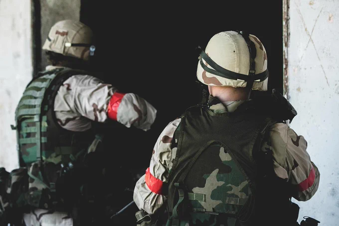 Two soldiers in camouflage gear and helmets enter a dark room, each wearing red armbands on their sleeves